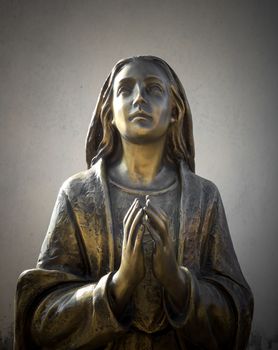 Holy Mary statue bronze that prays with folded hands, in the background a gray wall. It can be used for events, concepts and backgrounds.