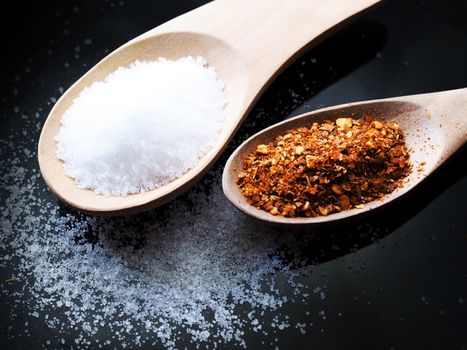 Dried cayenne pepper and salt in a wooden spoon on black background.