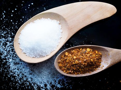 Dried cayenne pepper and salt in a wooden spoon on black background.
