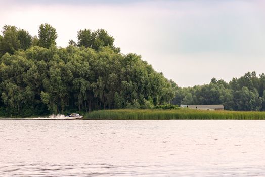 Little house close to the Dnieper river in Kiev, Ukraine, at the beginning of spring, under a cloudy sky. A black and white speedboat passes in front of the tender green trees.