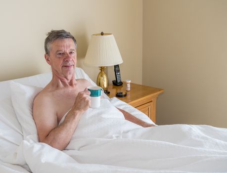 Senior retired caucasian man lying in adjustable bed on incline. He has a cup of coffee and is smiling towards the viewer