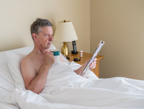 Senior retired caucasian man lying in adjustable bed on incline. He is reading from magazine with cup of coffee