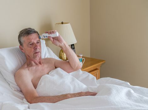 Senior retired caucasian man lying in adjustable bed on incline. He is checking his temperature for fever or signs of virus