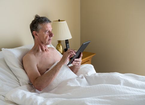 Senior retired caucasian man lying in adjustable bed on incline. He is reading and scrolling on digital tablet