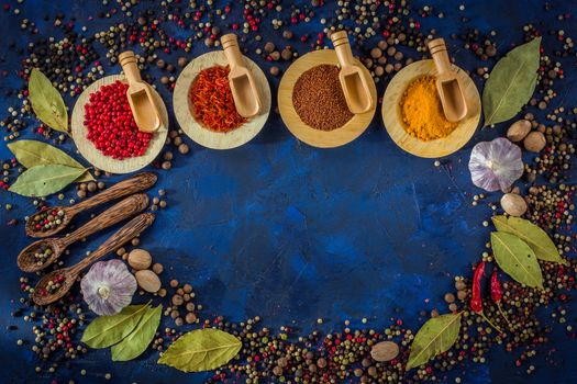 Assortment of colorful spices in the wooden bowls with spoons on a dark blue background. Curry, saffron, pink peppercorns, star anise, pepper, hot chili, garlic, bay leaf, nutmeg. Top view. Copy space.