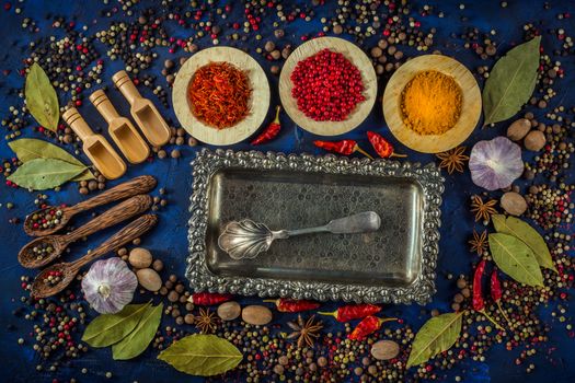 Assortment of colorful spices in the wooden bowls with spoons on a dark blue background. Curry, saffron, pink peppercorns, star anise, pepper, garlic, bay leaf and nutmeg with vintage silver plated tray with silver spoon. Top view. 