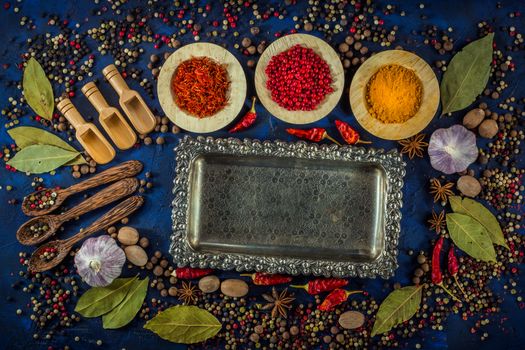 Assortment of colorful spices in the wooden bowls with spoons on a dark blue background. Curry, saffron, pink peppercorns, star anise, pepper, hot chili, garlic, bay leaf and nutmeg with vintage silver plated tray. Top view. 