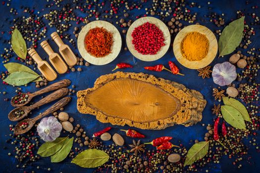 Assortment of colorful spices in the wooden bowls with spoons on a dark blue background. Curry, saffron, pink peppercorns, star anise, pepper, hot chili, garlic, bay leaf and nutmeg with cutting board. Top view. 