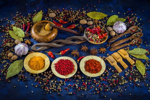 Various fragrant spices and herbs on a dark blue background. Spicy background with pepper mix and other spices. Colorful spices for cooking with vintage handmade wooden spoon and bowl. Top view, flat-lay.