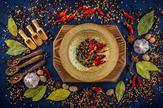 Assortment of colorful spices on a dark blue background. Spicy background with pepper mix on a ceramic plate. Colorful spices for cooking with handmade wooden spoon and bowl. Top view, flat-lay.