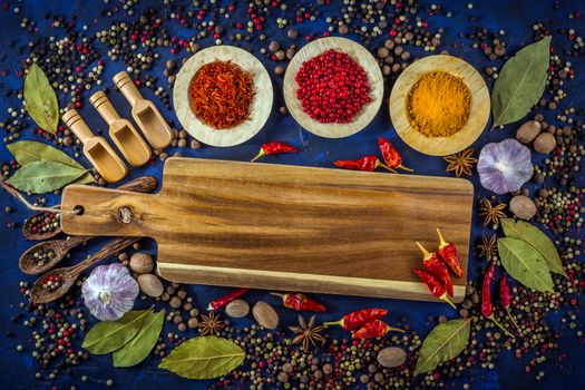 Assortment of colorful spices in the wooden bowls with spoons on a dark blue background. Curry, saffron, pink peppercorns, star anise, pepper, hot chili, garlic, bay leaf and nutmeg with cutting board. Top view. 