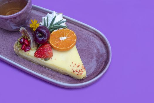 A serving of lemon tart topped with mixed fresh fruit with a cup of green tea on a purple ceramic plate.