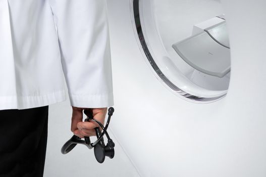 A doctor holding a black stethoscope standing in front of a CT scanner.
