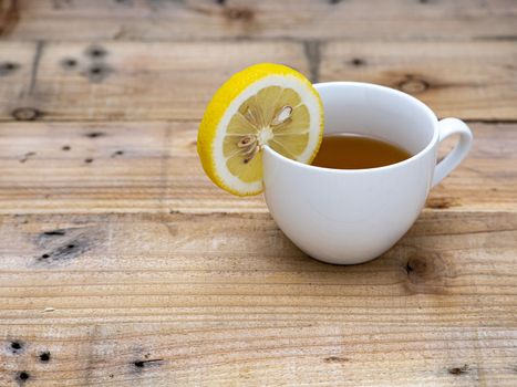 A cup of tea with a slice of lemon on its brim. Light brown wooden background.