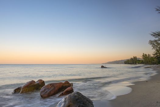 Sea waves splashing on the rocks at the sandy beach in the early morning with orange rays appearing in the horizon.