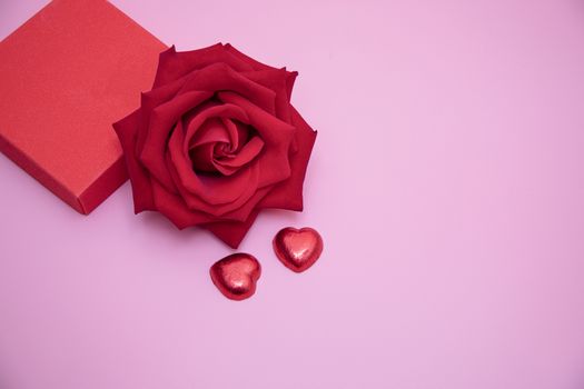 Closeup of a red rose with two chocolate candy hearts and a red gift box on pink background. Valentine's, anniversary, wedding concept.