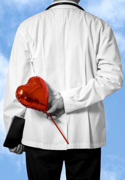 A male doctor in white coat holding a red balloon behind his back. Blue sky with white clouds background. Selective focus on red baloon with limited color. Doctor and caring concept.