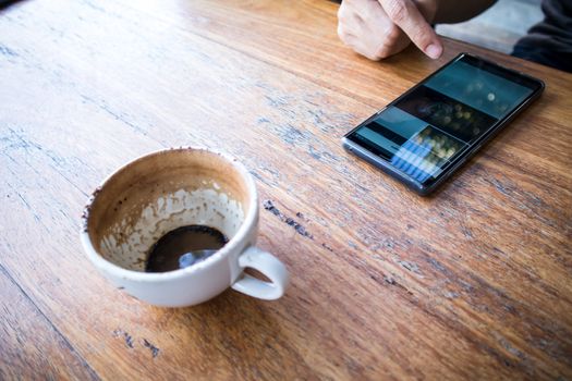 Close up of a right hand using smartphone with a Finished Empty Cup of Coffee on a brown wooden table. view from above