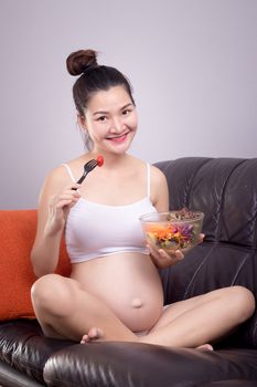 Pregnant nutrition healthy concept. Happy young beautiful Asian pregnant woman eat salad from salad bowl on sofa with smiley face. Health care with relax at home.Beautiful Asia female model in her 20s
