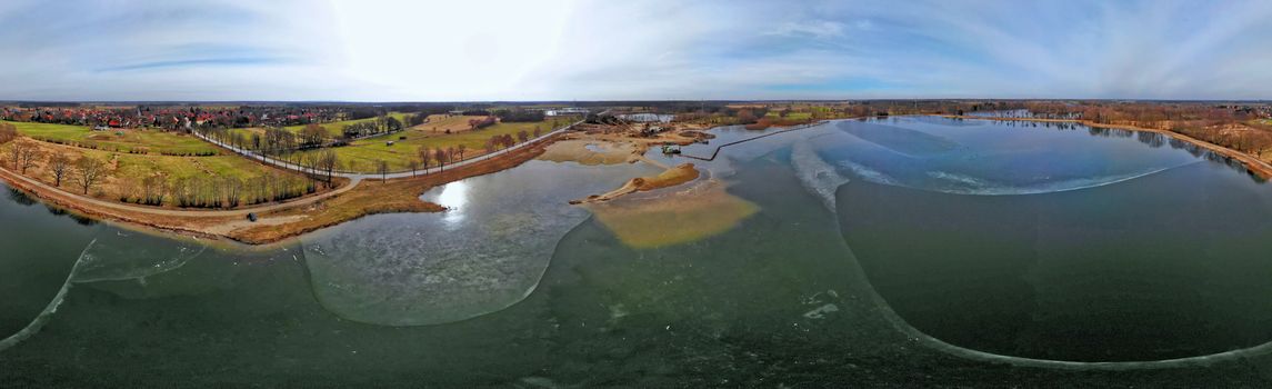 Composite panorama of aerial photographs and aerial photos of a wet quarry for gravel and sand with an almost frozen lake and large machines for excavation and processing, made with drone