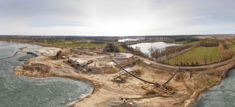 Composite panorama of aerial photographs and aerial photos of a wet quarry for gravel and sand with an almost frozen lake and large machines for excavation and processing, made with drone