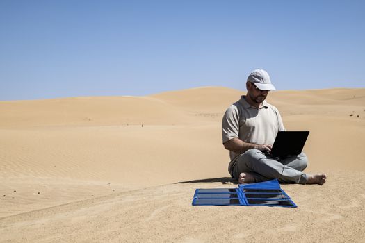 Adult (man) using his laptop in the Middle of the dunes by clear skies and using portable flexible solar panels and charger