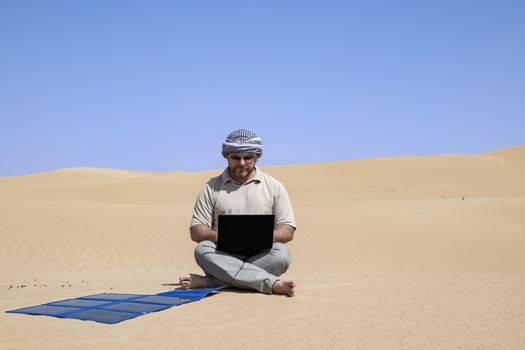 Man (adult) using his computer charging with flexible solar panels in the middle of the dunes.
