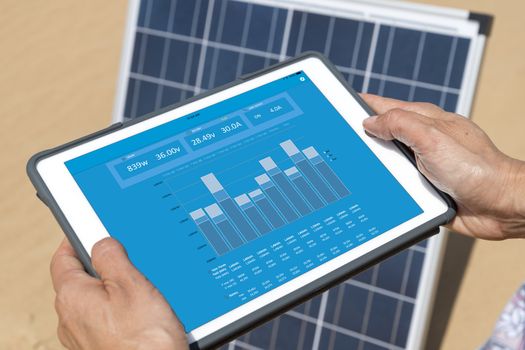 Woman hands on electronic tablet controlling solar panel with an app (blue screen with statistics and graph of the energy production & consumption)
