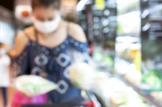 Blue background.  Asian woman making decision to choose products in Supermaket store. Defocus yong woman wear mask for shopping vegetable in supermarket store. New normal. 