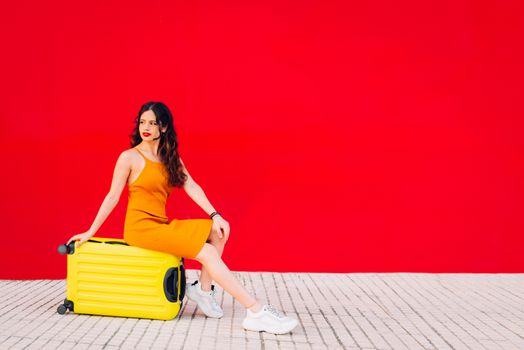 woman sitting on top of a yellow suitcase in front of a red wall.