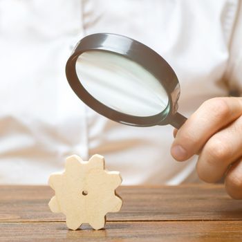 Businessman examines a wooden gear through a magnifying glass. A cog from a big business machine. Every little thing is important. study and analysis of business processes and subjects Research