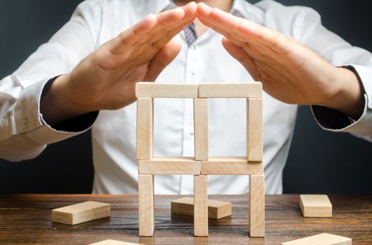 A man holds his hands in the symbol of the roof over the building of dominoes. Creating a business, startup. company protection, support for small and medium-sized businesses. Insurance assistance