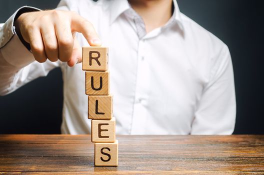 A man holds a tower of blocks with the word Rules from falling. Setting clear rule and restrictions. Leadership and discipline. Authoritarianism, tight control framework. Norms and laws in society