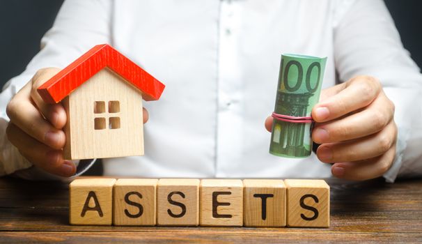 A man holds a house and a bundle roll of Euro over the word Assets. Declaration income and taxation, audit of ownership of real estate and cash. Reliable investment. Property tax