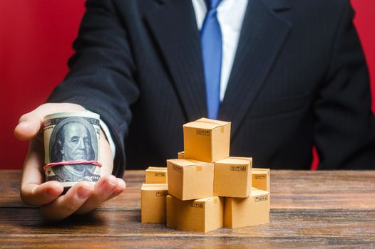 A businessman holds out a bundle of dollars near a pile of boxes. Trade and exchange goods, offers for cooperation. Profit, Superprofits. Investments financing in production, taxes, income revenues