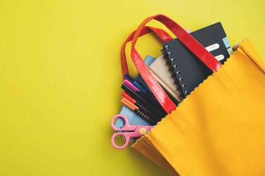 Learning supplies with notebooks, coloured pens and scissors in yellow bag on yellow background with copy space for education and back to school concept

