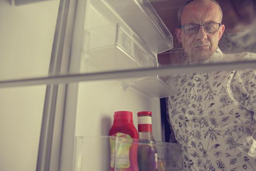 Senior man opening a refrigerator with little food. Selective focus. Hungry man looking for a late night meal in an empty fridge.