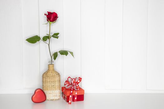 A single beautiflul red rose in a bamboo vase with a red gift box and a red paper mache heart. White painted teak wood background.