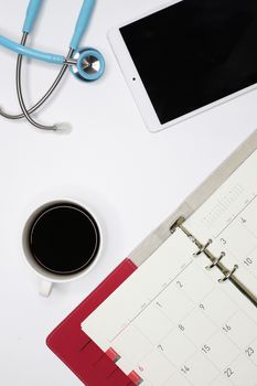 a top view image of a doctor's clean desk with an appointment book, a tablet, a cup of coffee, and a stethoscope