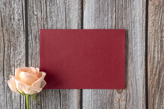 A piece of bright red paper with a single peach color rose on brown wooden background. Mock up invitation card. Top view. Flat design.