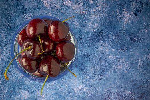 Fresh dark red cherries in clear glass of water on blue and white acrylic paint background. Closeup, top view.