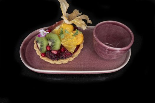 A piece of mixed fruit lemon tart on a purple ceramic plate with a tea cup. Black background.