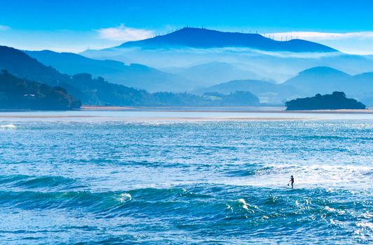 Beautiful landscape with foggy blue hills, ocean, and man on a surfboard