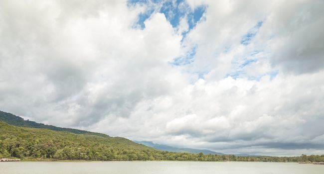 A panoramic view of a large water reservior surrounding by small hills under gathering rainy clouds and some blue sky.