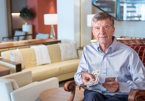 Senior caucasian man holding some US dollar bills as if handing them to the viewer. Composite into upmarket lounge of hotel or cruise ship