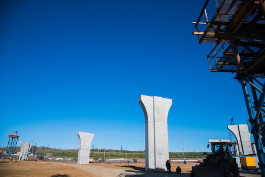 Construction of the interchange of a road bridge across the river. Builders and construction equipment on the construction site. The support of the bridge against the blue sky.