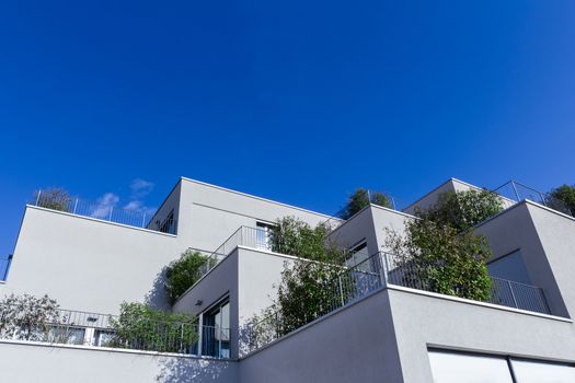 Grey apartment building with modern architecture and large balconies with green plants. Behind a background of blue sky. Bottom view.