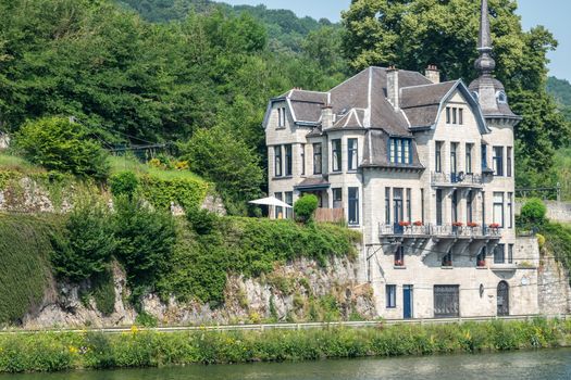 Dinant, Belgium - June 26, 2019: Light beige stone historic mansion on left bank of River Meuse against wall of green foliage is called Villa Mouchenne, top restaurant of region.