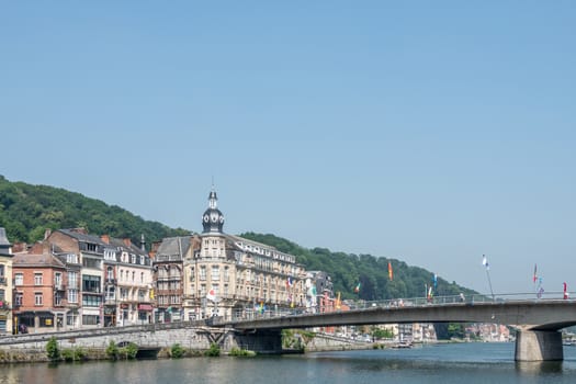 Dinant, Belgium - June 26, 2019: Charles de Gaule bridge spans Meuse River under blue sky. Left bank buildings with historic towered corner building. flags and green foliage.