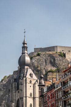 Dinant, Belgium - June 26, 2019: The citadel fort on cliff towers high above the spire of gray stone Notre Dame church under light blue sky. Housing builidngs in differnt colors.
